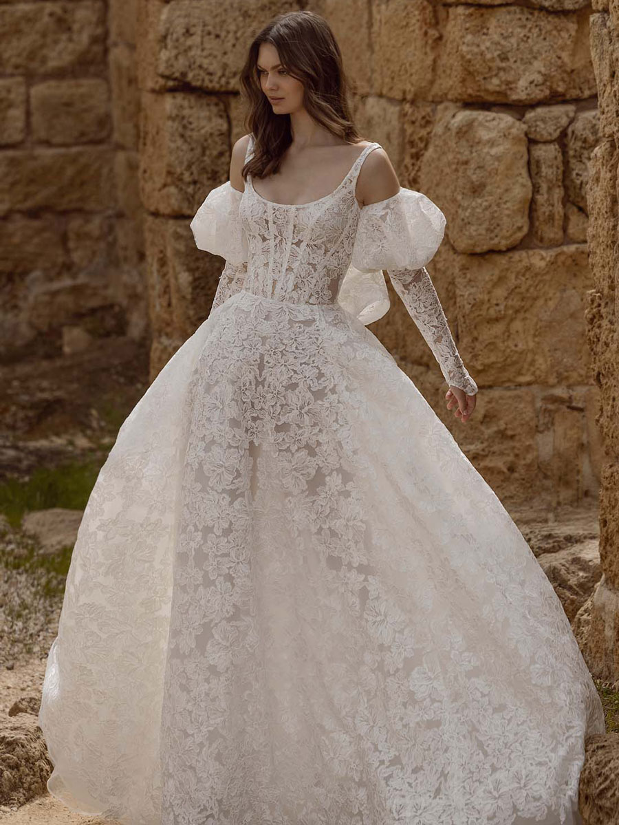 Love Couture Bridal Wedding Dresses. A woman in a sheer, lacy white wedding dresses gown from a bridal boutique, featuring puffed sleeves and cut-out shoulders, stands outdoors beside a stone wall. The designer dress boasts intricate floral patterns and a fitted bodice, complementing the flowing skirt. She has long, dark hair.