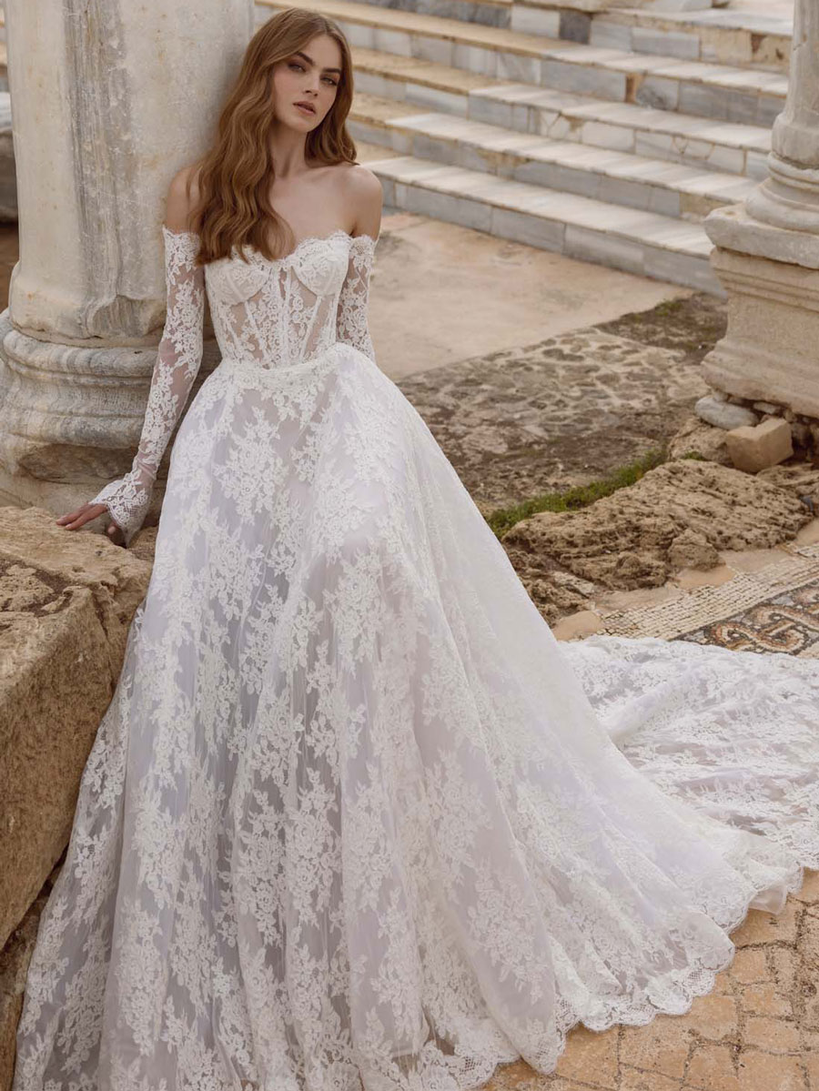 Love Couture Bridal Wedding Dresses. A woman in a long, white, off-the-shoulder lace wedding dresses from a renowned bridal boutique leans against ancient stone ruins. The dress has intricate floral lace details and a flowing train. She has long, wavy hair and appears contemplative against the historical backdrop of stone steps and columns.