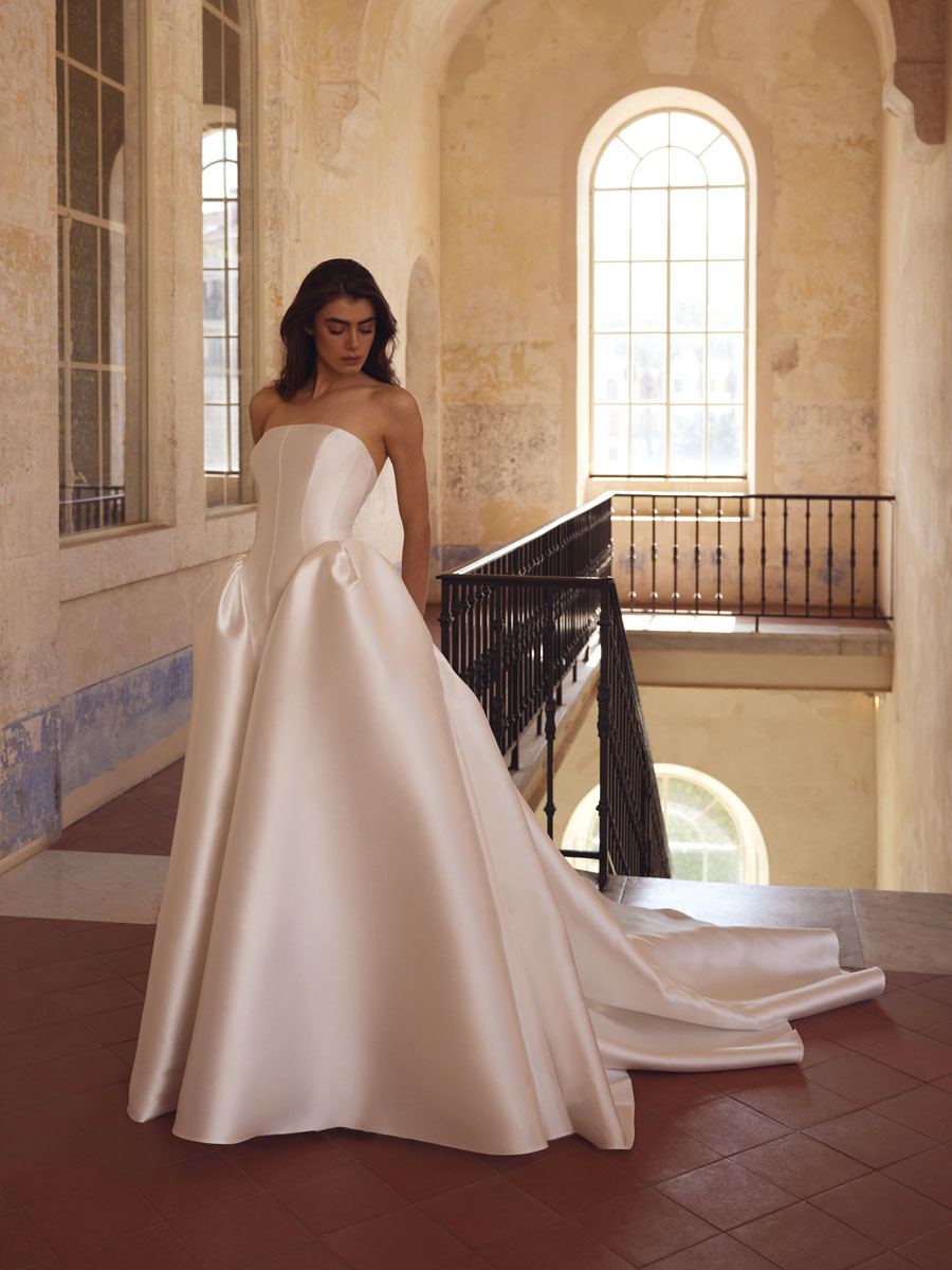 Love Couture Bridal Wedding Dresses. Woman in white wedding dresses leans against ancient stone columns.